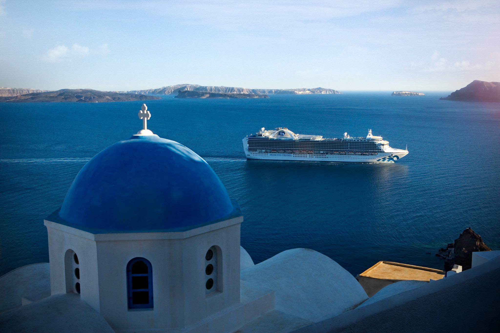 A Princess cruise ship sails through the ocean in Santorini Greece, with the iconic Blue Domed Church in the foreground.