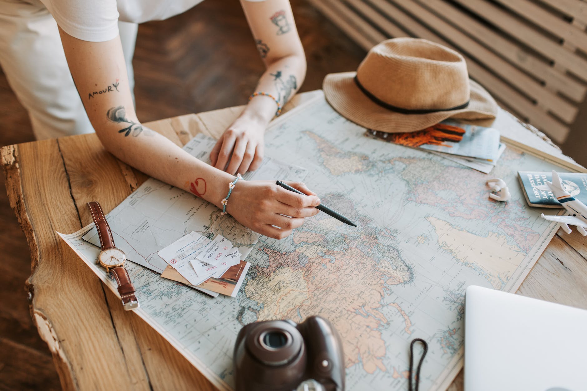 A woman holding a pen sits at a table covered with a large map of the world. There is a sunhat, a polaroid style camera, and some papers sitting out on top of the map.