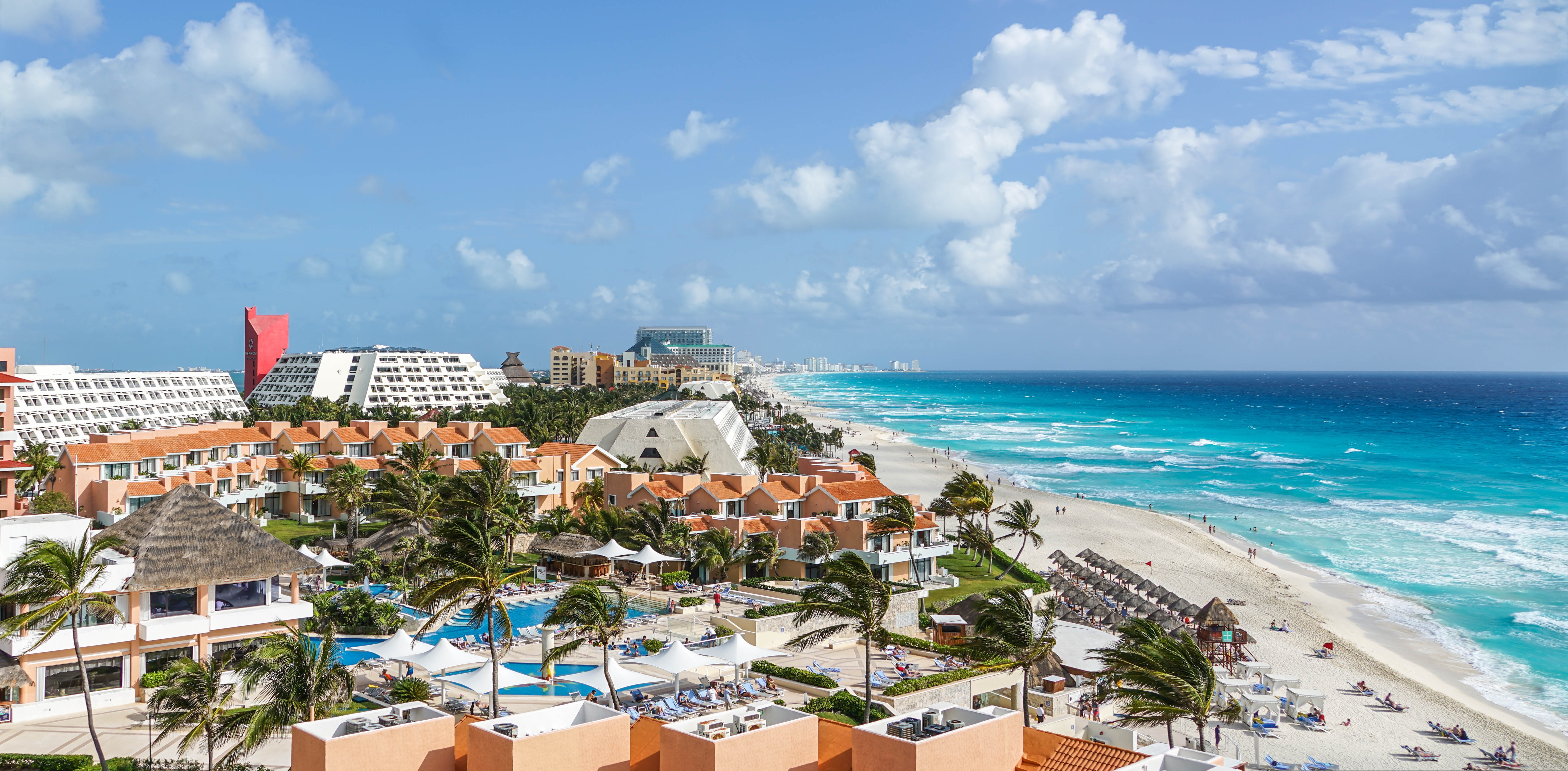 Birds eye view of Cape Cancun Beach in Mexico. On the left side of the image, there are many resort buildings with swimming pools. On the right side of the image, waves crash onto the beach where people are swimming, lounging in beach chairs and under umbrellas, and walking along the shore.