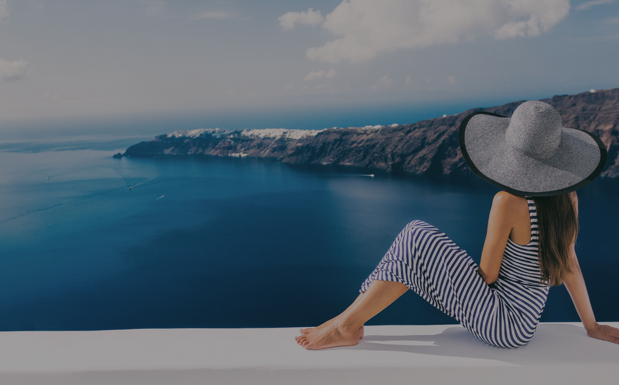A woman in a long blue and white striped dress and a gray sunhat lounges on a white ledge over looking the blue water and mountains in the background.